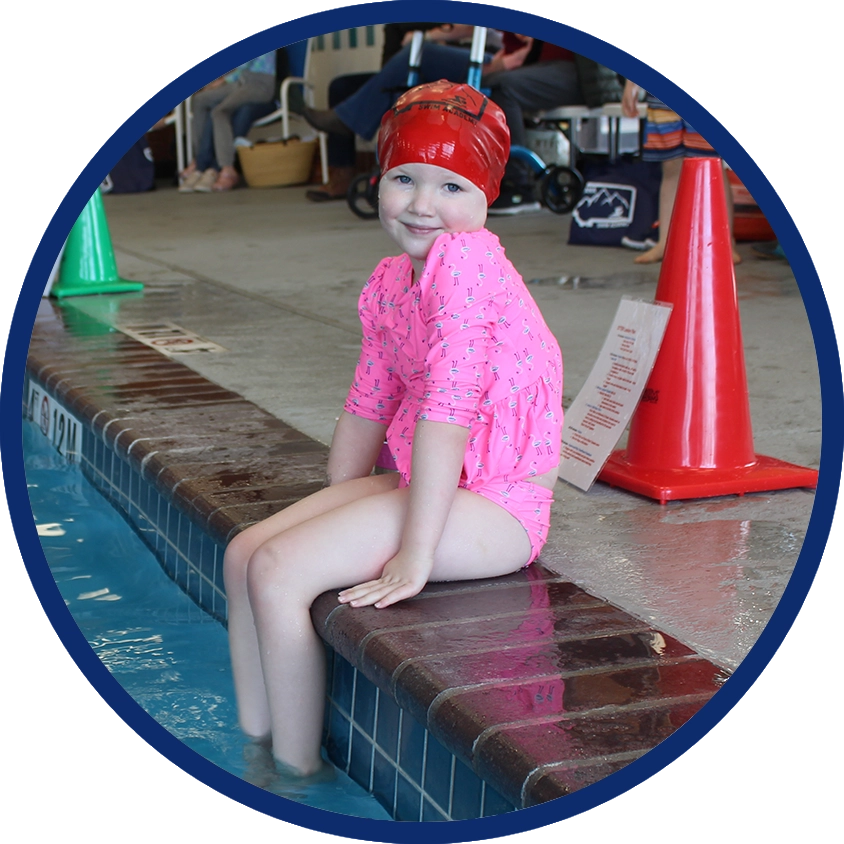 girl in red swim cap and pink bathing suit sitting at edge of pool and smiling at camera